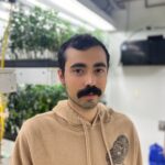 Daniel in a room of plants. He is wearing a tan hoodie.