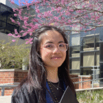 Annie standing outside in her cap and gown in front of a tree with pink blooms and a building behind.