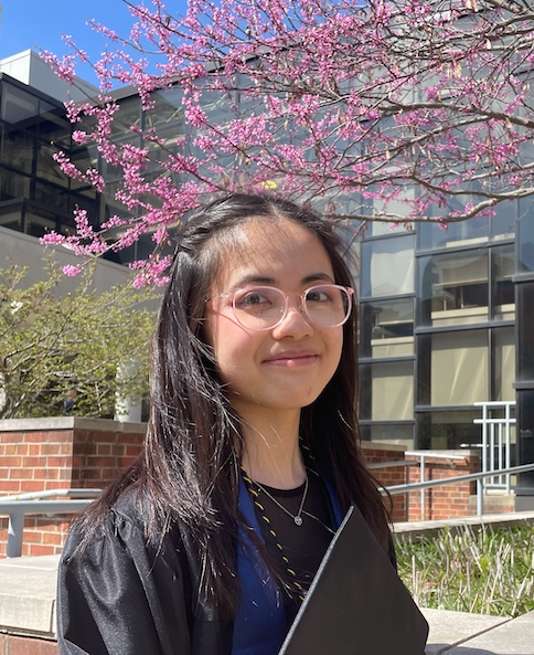 Annie standing outside in her cap and gown in front of a tree with pink blooms and a building behind.